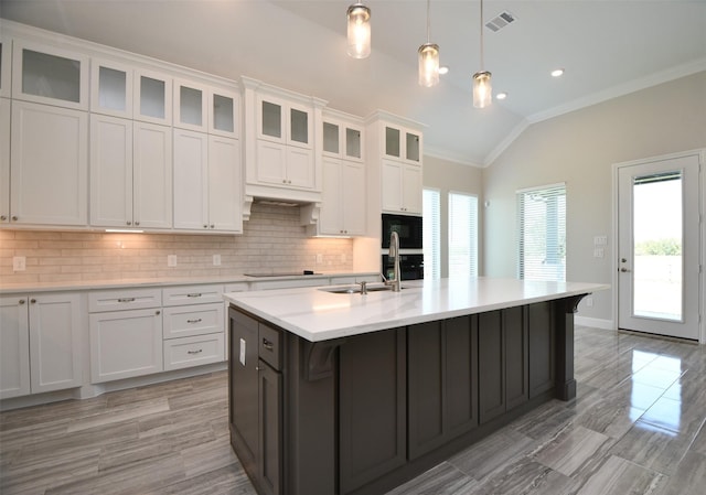 kitchen with ornamental molding, vaulted ceiling, a kitchen island with sink, pendant lighting, and white cabinets