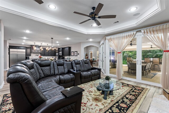 living room with a raised ceiling, ceiling fan with notable chandelier, and light wood-type flooring