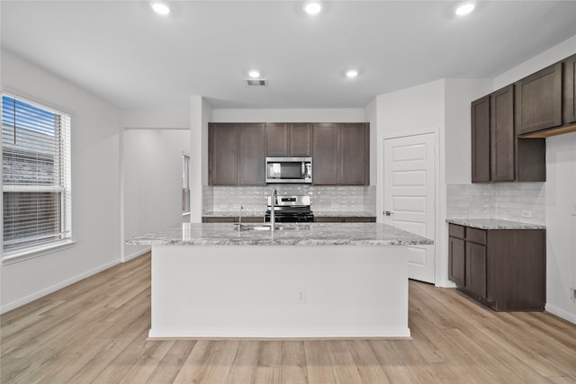 kitchen with stainless steel appliances, a kitchen island with sink, dark brown cabinets, and light wood-type flooring