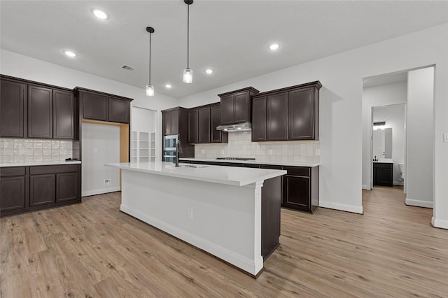 kitchen featuring decorative light fixtures, light wood-type flooring, backsplash, dark brown cabinets, and a center island with sink