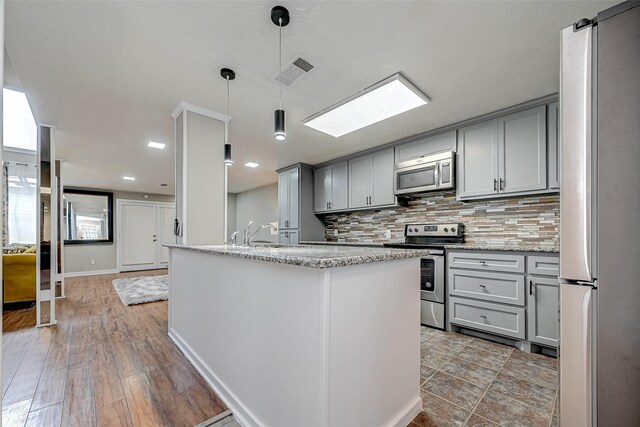 kitchen with tasteful backsplash, an island with sink, stainless steel appliances, pendant lighting, and gray cabinets