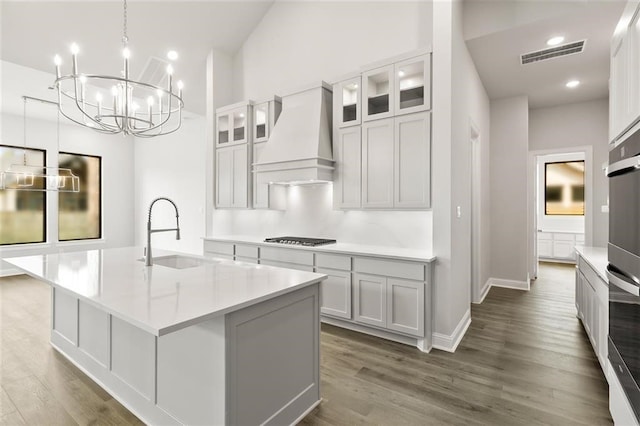 kitchen featuring sink, stainless steel appliances, premium range hood, decorative light fixtures, and a kitchen island with sink