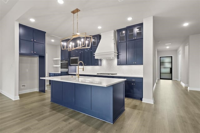 kitchen with stainless steel appliances, blue cabinetry, hanging light fixtures, a center island with sink, and custom range hood