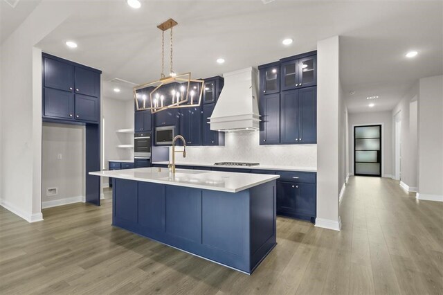 kitchen with stainless steel appliances, a kitchen island with sink, blue cabinets, hanging light fixtures, and custom range hood