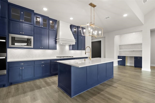 kitchen featuring a center island with sink, custom exhaust hood, sink, built in microwave, and blue cabinets