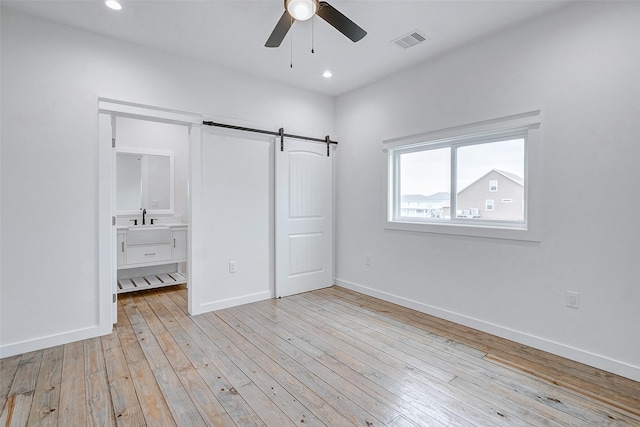 unfurnished bedroom with light wood-type flooring, ceiling fan, sink, a barn door, and connected bathroom