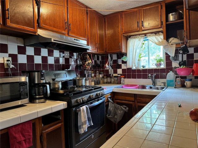 kitchen with tile counters, a sink, stainless steel appliances, under cabinet range hood, and backsplash