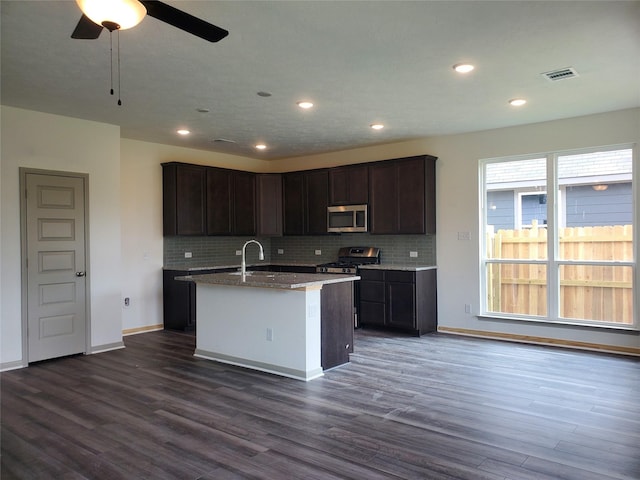 kitchen with dark wood-style flooring, stainless steel appliances, visible vents, backsplash, and a kitchen island with sink