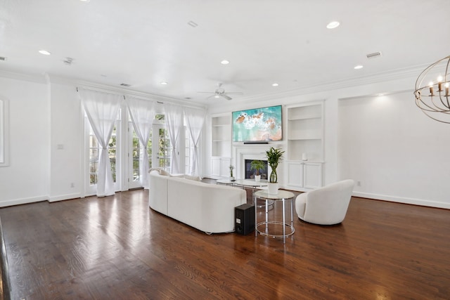 living room with built in shelves, dark hardwood / wood-style flooring, ceiling fan with notable chandelier, and ornamental molding