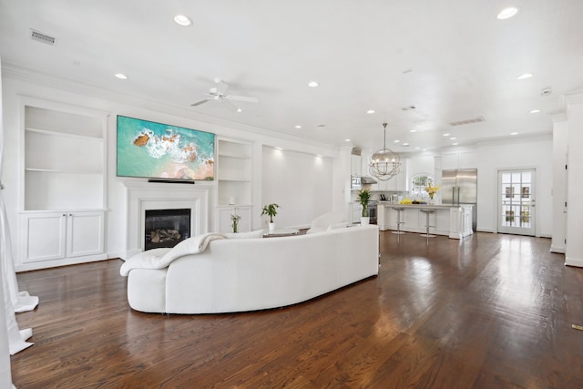 living room with built in shelves, ornamental molding, wood-type flooring, and ceiling fan with notable chandelier