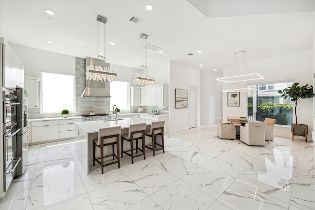 kitchen featuring wall chimney range hood, an island with sink, a healthy amount of sunlight, and white cabinets