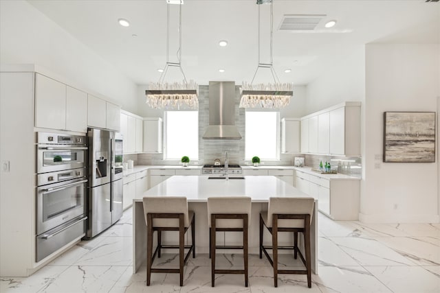 kitchen featuring wall chimney range hood, stainless steel appliances, an island with sink, decorative light fixtures, and backsplash