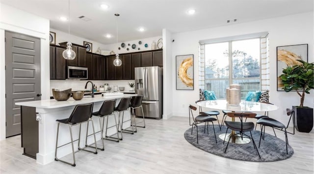 kitchen featuring stainless steel appliances, an island with sink, dark brown cabinets, pendant lighting, and tasteful backsplash