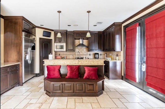 kitchen with dark brown cabinetry, built in appliances, hanging light fixtures, and wall chimney range hood