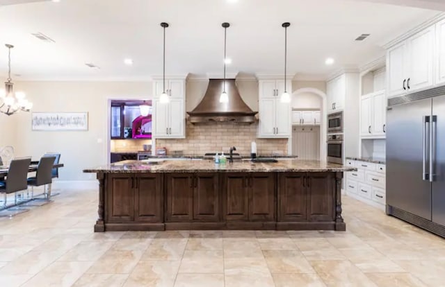 kitchen featuring light stone countertops, built in appliances, hanging light fixtures, wall chimney exhaust hood, and white cabinetry