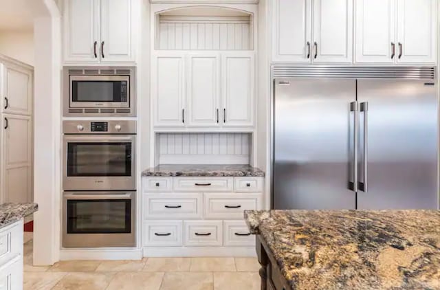 kitchen with dark stone counters, built in appliances, white cabinets, and light tile flooring