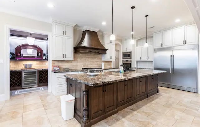 kitchen featuring tasteful backsplash, built in appliances, an island with sink, white cabinets, and premium range hood