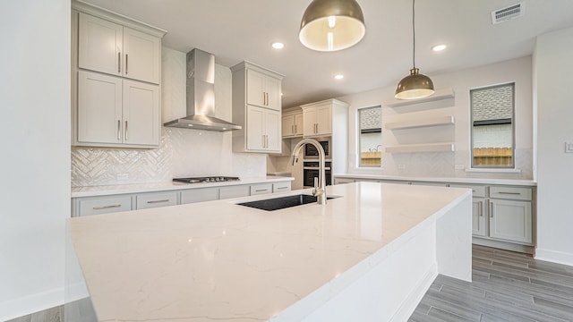 kitchen featuring gray cabinetry, light stone countertops, wall chimney exhaust hood, pendant lighting, and a kitchen island with sink