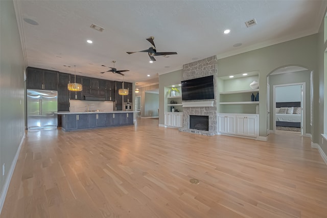 unfurnished living room with ornamental molding, light wood-type flooring, built in shelves, a stone fireplace, and ceiling fan