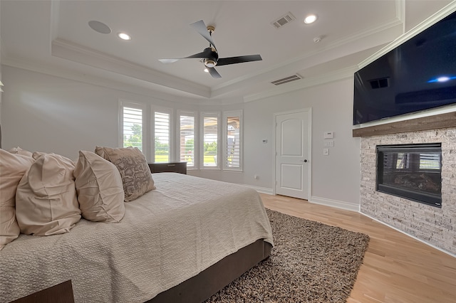 bedroom featuring a stone fireplace, ceiling fan, light hardwood / wood-style flooring, a raised ceiling, and ornamental molding