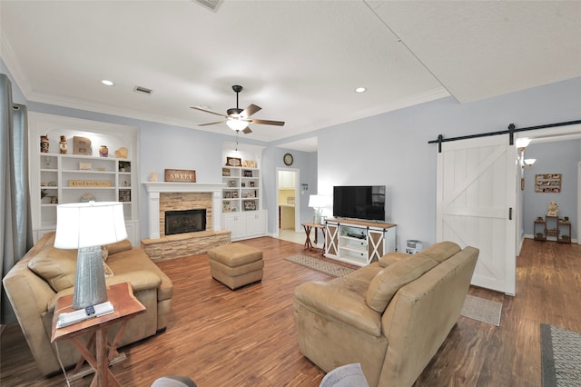 living room featuring wood-type flooring, ceiling fan, crown molding, a barn door, and a stone fireplace