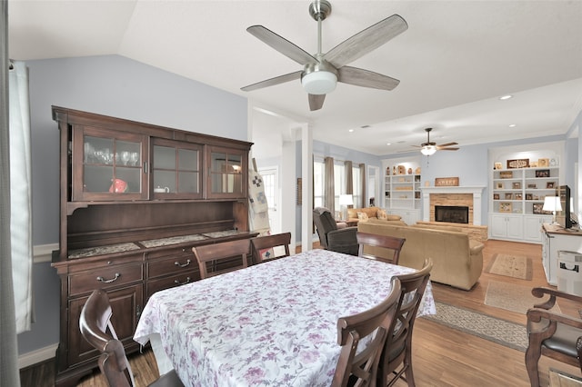 dining area featuring built in features, vaulted ceiling, a stone fireplace, light wood-type flooring, and ceiling fan