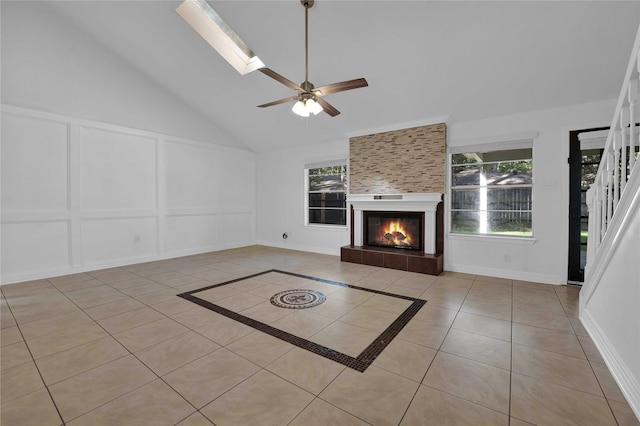 unfurnished living room with high vaulted ceiling, a skylight, a tiled fireplace, light tile patterned floors, and ceiling fan