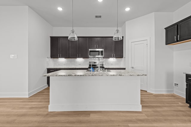 kitchen with light stone counters, an island with sink, decorative light fixtures, and stainless steel appliances