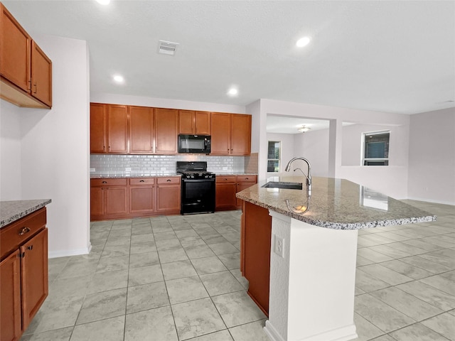 kitchen featuring an island with sink, sink, backsplash, black appliances, and light stone countertops