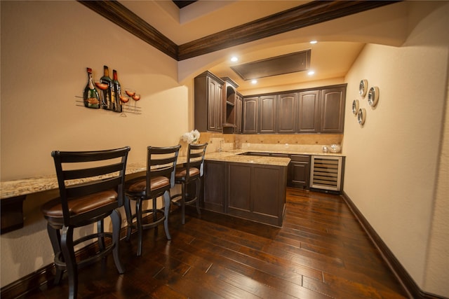 bar featuring dark brown cabinetry, light stone countertops, beverage cooler, dark wood-type flooring, and ornamental molding