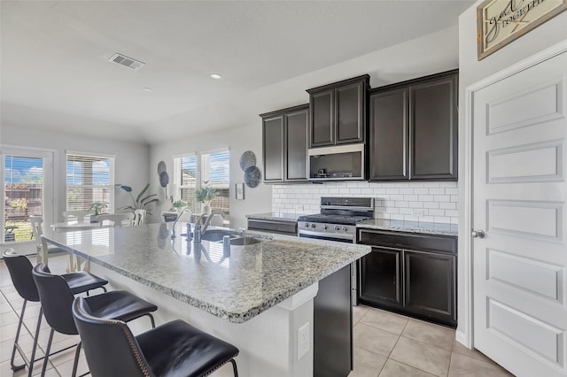 kitchen with sink, decorative backsplash, an island with sink, appliances with stainless steel finishes, and a kitchen bar