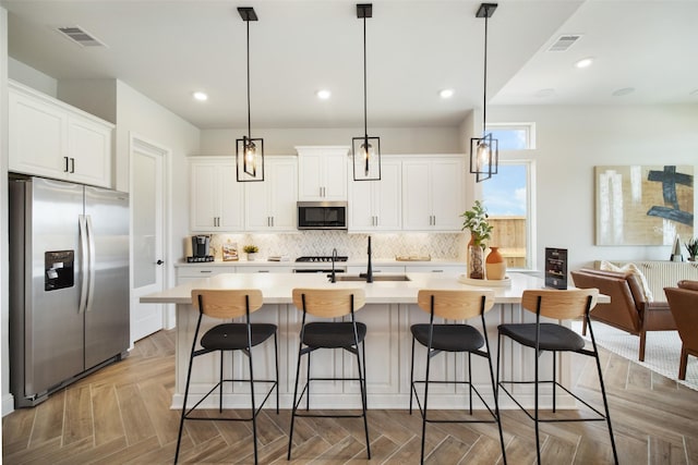 kitchen featuring light parquet floors, white cabinetry, an island with sink, stainless steel appliances, and hanging light fixtures