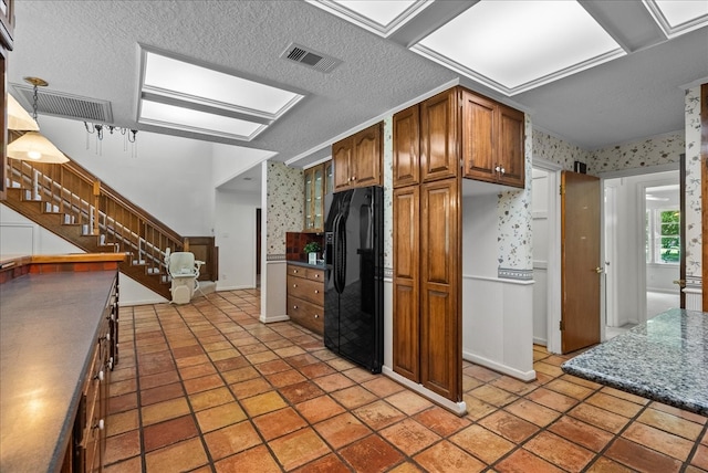 kitchen featuring a textured ceiling, black fridge with ice dispenser, visible vents, brown cabinetry, and wallpapered walls