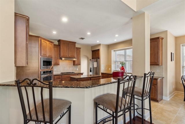 kitchen with kitchen peninsula, appliances with stainless steel finishes, a kitchen bar, decorative backsplash, and dark stone countertops