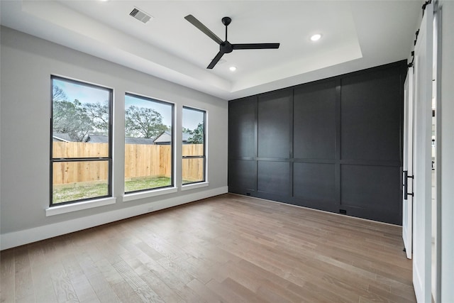 unfurnished room featuring ceiling fan, a tray ceiling, a barn door, and light wood-type flooring