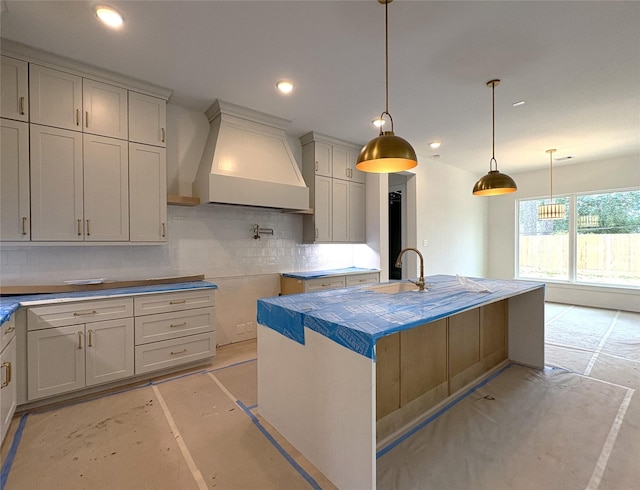 kitchen with premium range hood, hanging light fixtures, sink, backsplash, and an island with sink