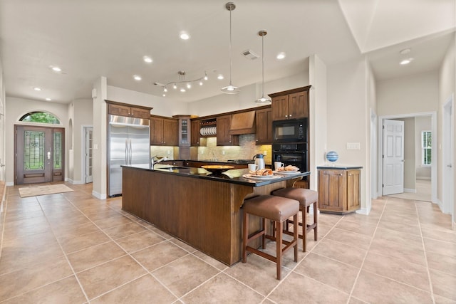 kitchen with light tile patterned floors, an island with sink, backsplash, custom range hood, and black appliances