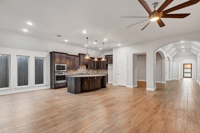 kitchen featuring light wood-type flooring, tasteful backsplash, stainless steel appliances, a center island with sink, and hanging light fixtures