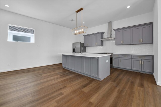 kitchen featuring wall chimney range hood, an island with sink, gray cabinets, dark wood-type flooring, and appliances with stainless steel finishes