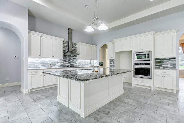 kitchen featuring wall chimney exhaust hood, a center island with sink, backsplash, and appliances with stainless steel finishes