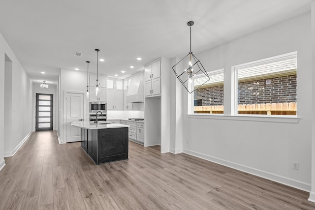 kitchen with custom exhaust hood, a center island with sink, sink, decorative light fixtures, and light hardwood / wood-style floors