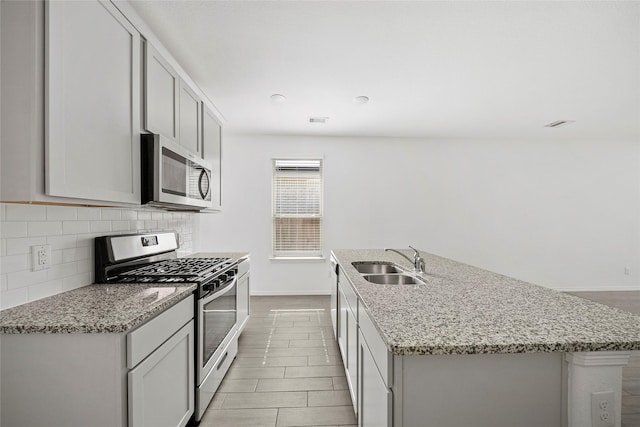 kitchen featuring appliances with stainless steel finishes, decorative backsplash, a kitchen island with sink, light stone countertops, and sink