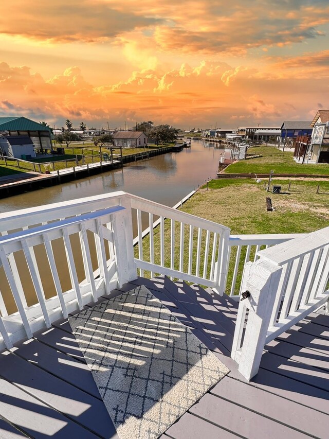 deck at dusk with a dock, a water view, and a lawn