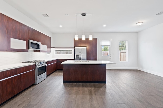 kitchen with appliances with stainless steel finishes, tasteful backsplash, decorative light fixtures, dark hardwood / wood-style floors, and an island with sink