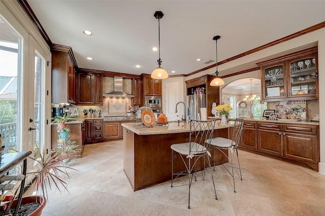 kitchen featuring wall chimney exhaust hood, decorative light fixtures, appliances with stainless steel finishes, an island with sink, and light stone countertops