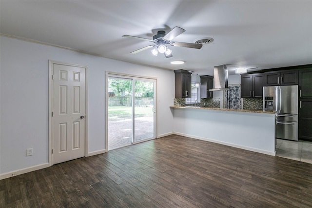 kitchen with dark wood-type flooring, wall chimney range hood, stainless steel fridge, tasteful backsplash, and kitchen peninsula