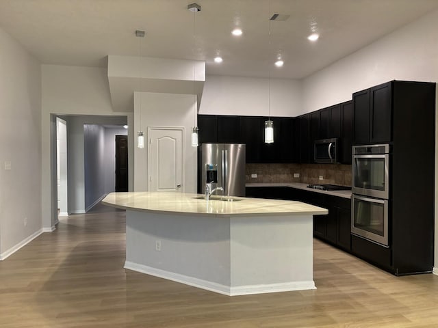 kitchen with backsplash, a kitchen island with sink, a towering ceiling, decorative light fixtures, and stainless steel appliances