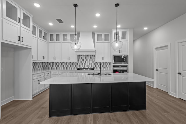 kitchen with premium range hood, white cabinets, sink, an island with sink, and appliances with stainless steel finishes