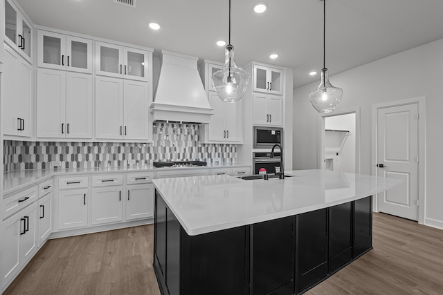 kitchen with custom range hood, sink, decorative light fixtures, white cabinets, and an island with sink