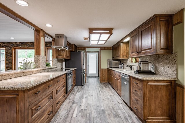 kitchen with decorative backsplash, island exhaust hood, light stone counters, appliances with stainless steel finishes, and light hardwood / wood-style flooring